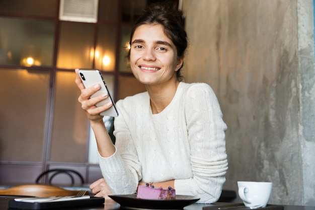 Immagine di bella giovane donna seduta al caffè a bere il caffè al chiuso utilizzando il telefono cellulare in chat.