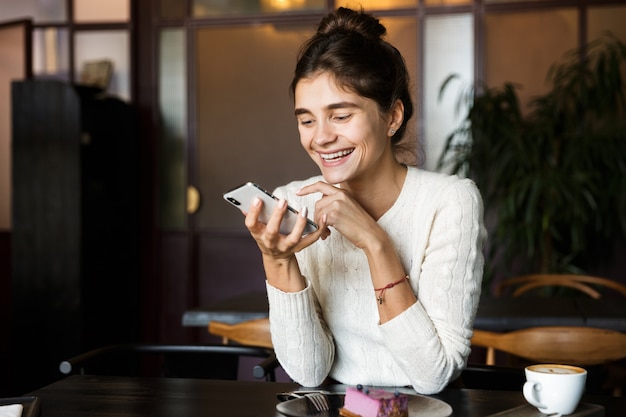 Immagine di bella giovane donna seduta al caffè a bere il caffè al chiuso utilizzando il telefono cellulare in chat.