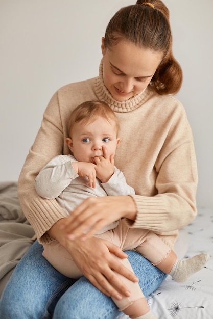 Immagine di donna dai capelli scuri con coda di cavallo che indossa un maglione beige che tiene la bambina in mano mentre è seduta sul letto madre che guarda la figlia giorni in congedo di maternità