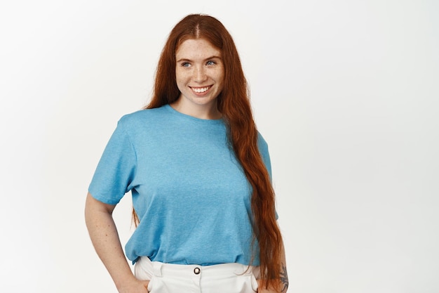 Immagine di donna sinuosa carina con lunghi capelli rossi naturali, lentiggini sul viso, sorridente e guardando da parte lo striscione con un affare promozionale di vendita, in piedi su sfondo bianco.