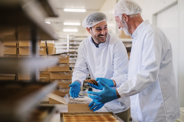 Immagine di due impiegati maschii della fabbrica dell'alimento in vestiti sterili che imballano i biscotti fatti freschi e che si divertono.