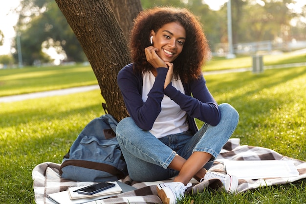 Immagine di felice giovane donna africana seduti all'aperto nel parco ascoltando musica con gli auricolari.