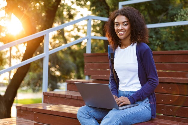 Immagine di felice giovane donna africana seduti all'aperto nel parco sui gradini utilizzando il computer portatile.