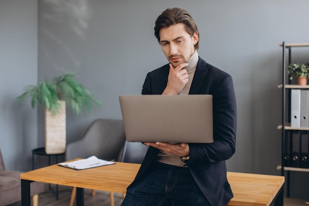 Immagine di giovane uomo d'affari allegro in giacca e cravatta che tiene e utilizza il laptop nell'ufficio moderno