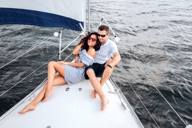 Immagine di giovani e belle coppie che si siedono e che si trovano sulla prua dell'yacht. Indossano occhiali da sole. . Sorrisi bruna. Si siede con le gambe incrociate e guarda avanti.