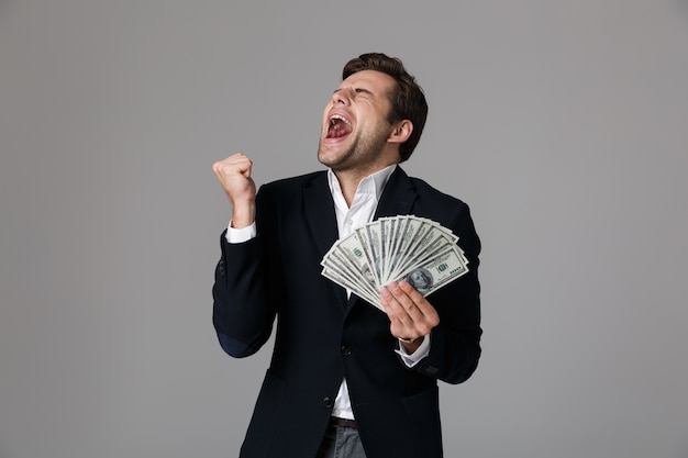 Immagine di happy businessman 30s in suit sorridente e holding fan di denaro in banconote in dollari, isolato sopra il muro grigio
