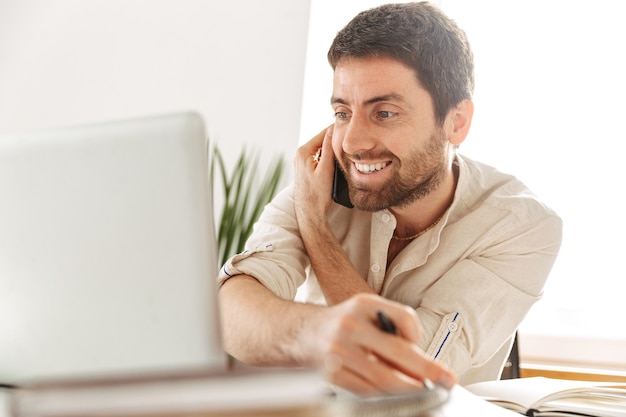 Immagine di happy businessman 30s indossa una camicia bianca parlando al telefono cellulare, mentre è seduto a tavola in un ufficio moderno