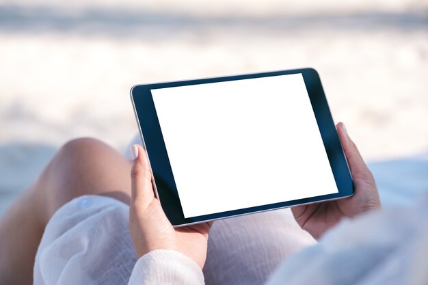 Immagine di mockup di una donna che tiene un tablet pc nero con schermo desktop vuoto mentre si sdraia sulla sedia a sdraio sulla spiaggia