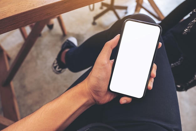 Immagine di mockup vista dall'alto della mano di un uomo che tiene il telefono cellulare bianco con lo schermo del desktop vuoto sulla coscia mentre è seduto nella caffetteria