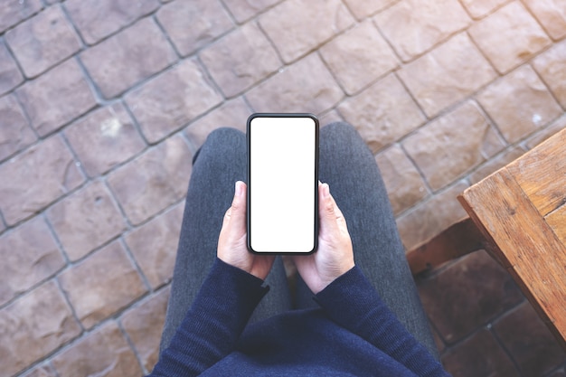 Immagine di mockup vista dall'alto delle mani che tengono il telefono cellulare nero con schermo bianco vuoto mentre è seduto all'aperto