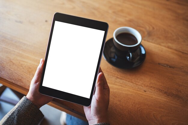 Immagine di mockup vista dall'alto delle mani che tengono un tablet pc nero con schermo bianco vuoto con tazza di caffè sul tavolo di legno