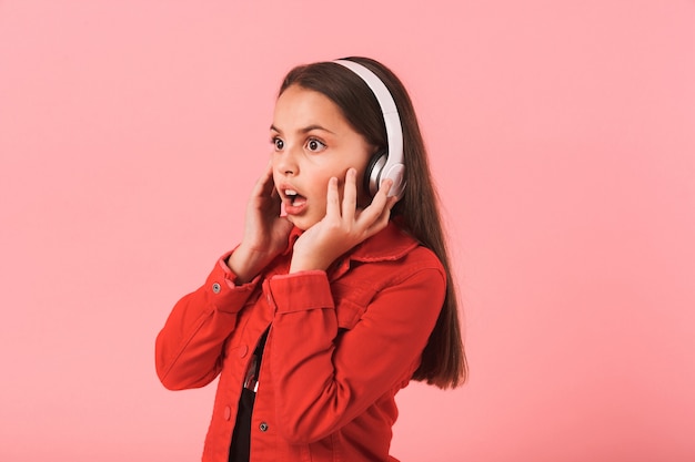 Immagine di musica d'ascolto della bella bambina scioccata con gli auricolari isolati sopra la parete rosa.