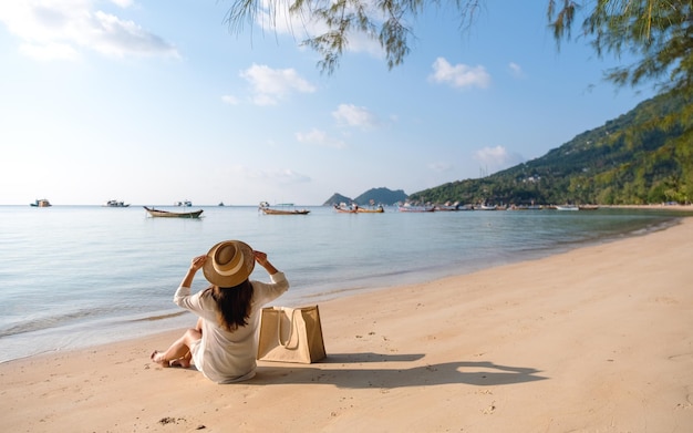 Immagine di retrovisione di una donna con cappello e borsa seduta sulla spiaggia con sfondo azzurro del cielo