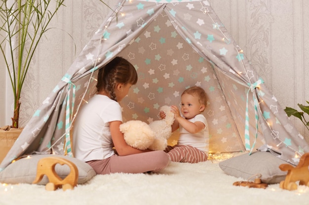 Immagine di sorelline che giocano nella tenda pipì con morbido orsacchiotto bambino che guarda il giocattolo con grande interesse fratello seduto sul pavimento su un morbido tappeto che trascorre del tempo insieme