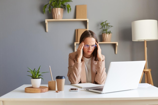 Immagine di triste disperazione stanca donna caucasica malata che indossa una giacca beige in posa in ufficio lavorando su un notebook guardando lo schermo faccia accigliata che ha problemi o soffre di mal di testa