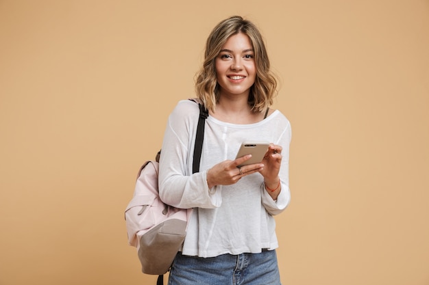 Immagine di un'adolescente bionda che indossa uno zaino usando il cellulare e sorridente isolata su un muro beige