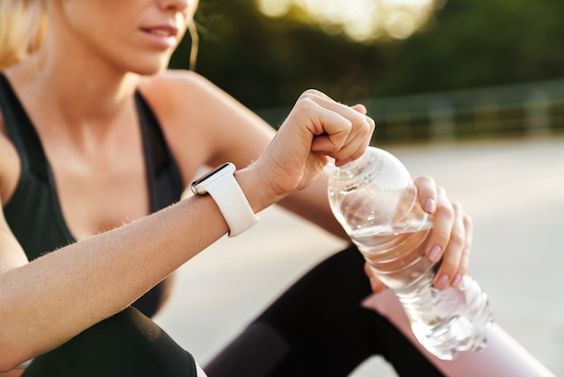 Immagine di un'adorabile sportiva in tuta da ginnastica che beve acqua durante l'allenamento sul tappetino fitness all'aperto