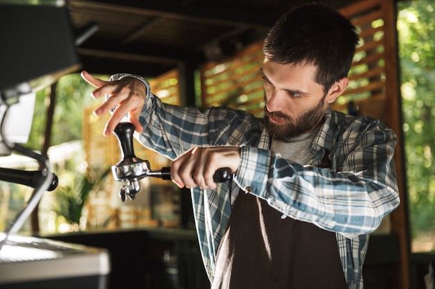 Immagine di un barista serio che indossa un grembiule che fa il caffè mentre lavora al bar o al caffè all'aperto