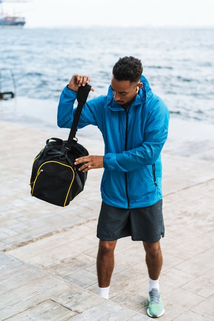 Immagine di un bel giovane africano sportivo uomo all'aperto in spiaggia mare a piedi con borsa ascoltando musica con gli auricolari