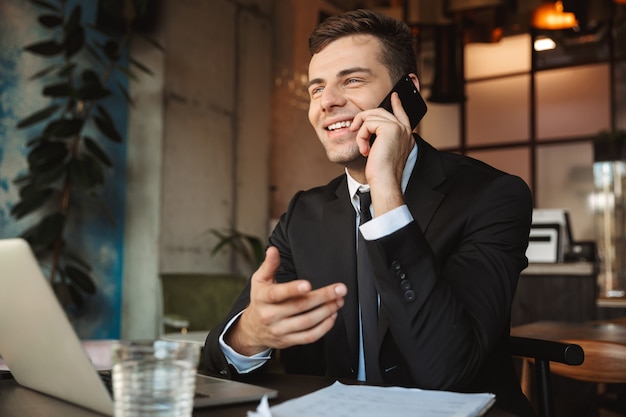 Immagine di un bel giovane imprenditore felice seduto in un caffè utilizzando il computer portatile parlando al telefono cellulare.