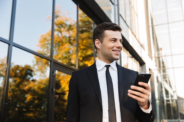 Immagine di un bel giovane uomo d'affari felice camminare all'aperto vicino al centro business utilizzando il telefono cellulare.