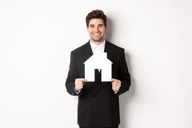Immagine di un bell'agente immobiliare in abito nero che mostra il maket domestico, sorride alla telecamera, in piedi su sfondo bianco.