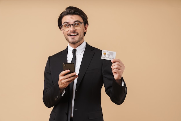 Immagine di un bell'uomo d'affari in abito formale con cellulare e carta di credito isolato su un muro beige