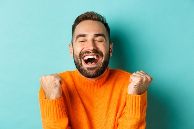 Immagine di un bell'uomo sollevato che si sente soddisfatto, si rallegra della vittoria o del risultato, fa pompare il pugno e dice di sì, in piedi su sfondo turchese.