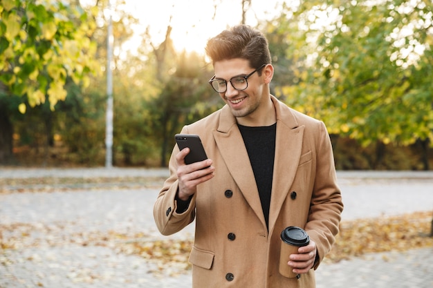 Immagine di un bell'uomo sorridente che indossa un cappotto digitando il cellulare e bevendo caffè da asporto mentre si cammina nel parco autunnale