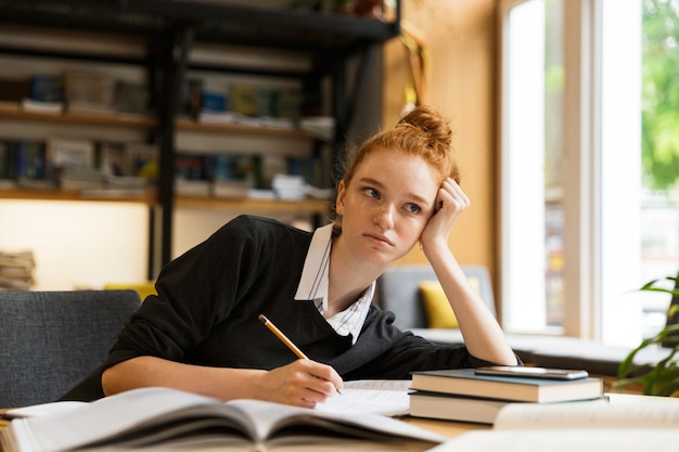 Immagine di un bellissimo studente che studia, mentre è seduto alla scrivania nella biblioteca del college con la parete dello scaffale