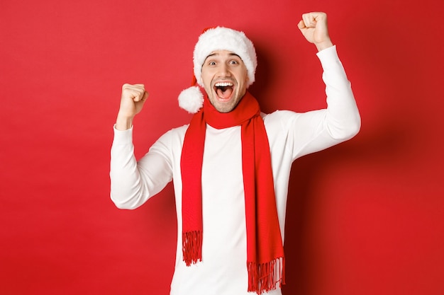 Immagine di un gioioso uomo caucasico con cappello e sciarpa da Babbo Natale che grida di gioia e alza le mani per celebrare ...
