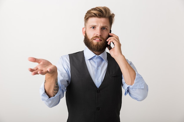 Immagine di un giovane barbuto serio confuso in piedi isolato sopra priorità bassa bianca della parete che parla dal telefono cellulare.