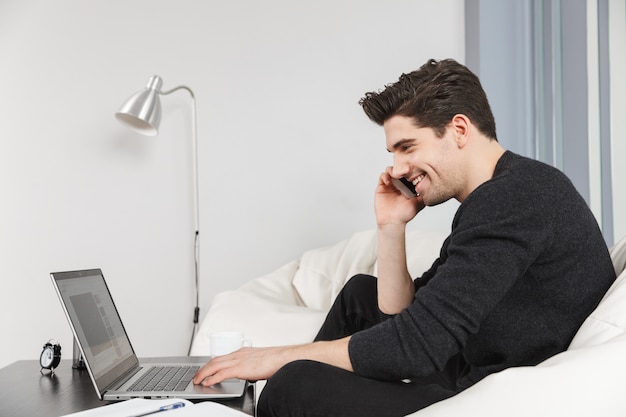 Immagine di un giovane bello felice a casa al chiuso utilizzando il computer portatile parlando dal telefono cellulare.