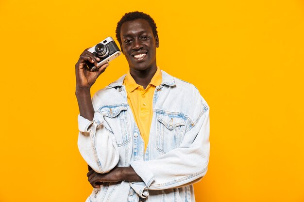 Immagine di un giovane ragazzo afroamericano in giacca di jeans che sorride e fotografa con una fotocamera retrò