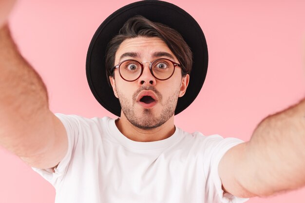 Immagine di un giovane sorpreso che indossa occhiali e cappello nero che scatta una foto selfie isolata