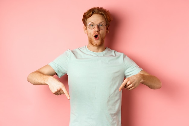 Immagine di un giovane stupito con i capelli e la barba rossi che indossano occhiali e maglietta che puntano le dita verso il basso...