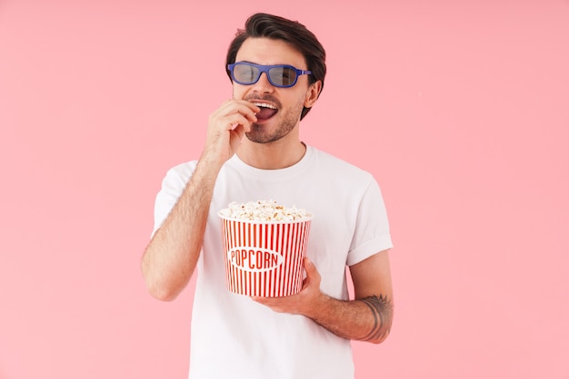 Immagine di un giovane uomo divertente che indossa occhiali 3D che mangia popcorn mentre guarda un film isolato