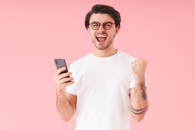 Immagine di un giovane uomo eccitato che indossa occhiali che tiene il cellulare e mostra il gesto del vincitore isolato