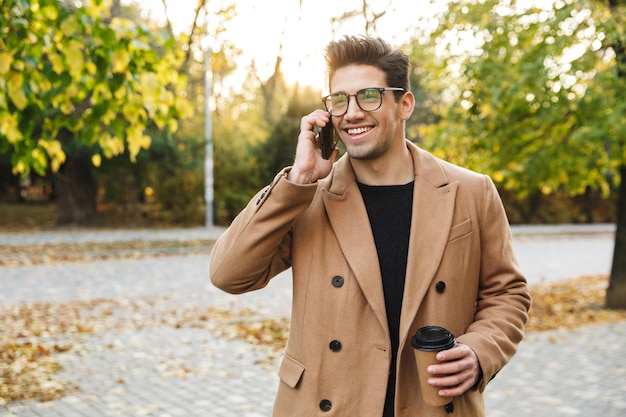 Immagine di un giovane uomo sorridente che indossa un cappotto che parla al cellulare e beve caffè da asporto mentre cammina nel parco autunnale