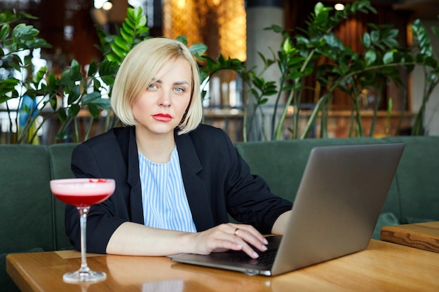 Immagine di un modello attraente ispirato che scrive al suo laptop trascorrendo del tempo da solo al bar con un bicchiere di cocktail sul tavolo essendo impegnato a indossare un abito