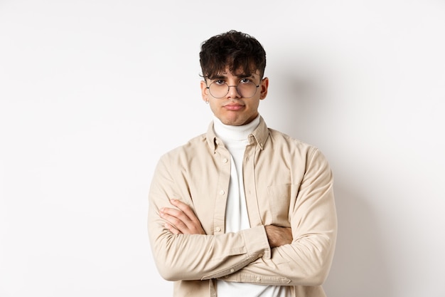 Immagine di un ragazzo alla moda hipster con gli occhiali che si sente annoiato, sembra non divertito con la faccia da poker, incrocia le braccia sul petto e sta riluttante contro il muro bianco.