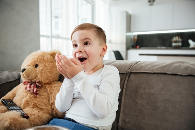 Immagine di un ragazzo sorpreso sul divano con un orsacchiotto a casa che guarda la TV mentre mangia patatine.