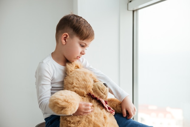 Immagine di un ragazzo triste vicino alla finestra con un orsacchiotto in attesa dei genitori a casa. Guardare oltre.