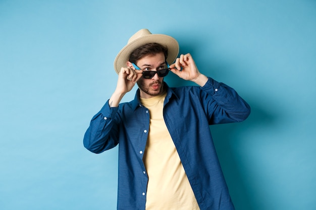 Immagine di un turista che guarda qualcosa di interessante, occhiali da sole da togliere, ad esempio wow e guarda da parte impressionato, in piedi su sfondo blu.