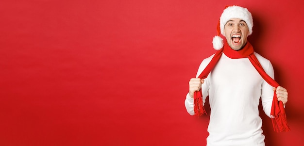 Immagine di un uomo adulto arrabbiato che odia il natale indossando sciarpa e cappello di Babbo Natale che urla angosciato in piedi matto ...