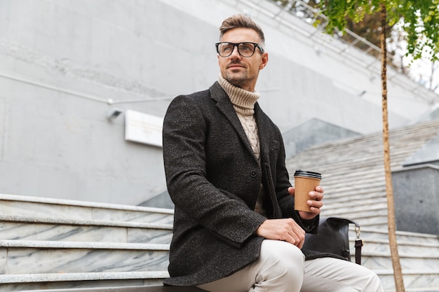 Immagine di un uomo alla moda degli anni '30 che indossa occhiali da vista, seduto sulle scale con caffè da asporto mentre passeggia per la strada della città