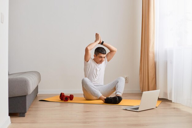 Immagine di un uomo atletico che fa yoga guardando il video di allenamento a casa in soggiorno durante il giorno le braccia alzate in un gesto di preghiera guardando lo schermo del laptop che si allena a casa