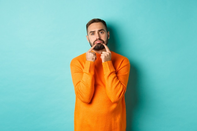 Immagine di un uomo barbuto cupo, che fa la faccia triste e si acciglia, in piedi sconvolto con un maglione arancione su sfondo turchese