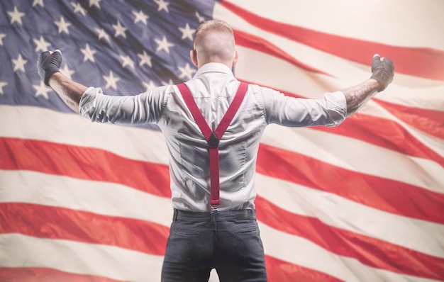 Immagine di un uomo brutale con una camicia bianca e bretelle rosse in piedi davanti alla bandiera americana. Il concetto di scazzottate. Tecnica mista