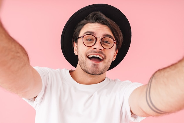 Immagine di un uomo caucasico che indossa occhiali e cappello che sorride mentre scatta una foto selfie isolata